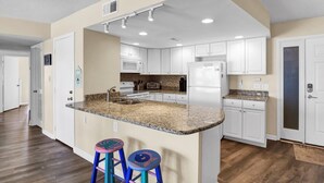 A modern kitchen with white cabinets, granite countertops, and stainless steel appliances. There are two stools at the island and a microwave above the sink. - Oceanfront Point Unit with 3 Bedrooms and Great Oceanviews (North Topsail Beach)