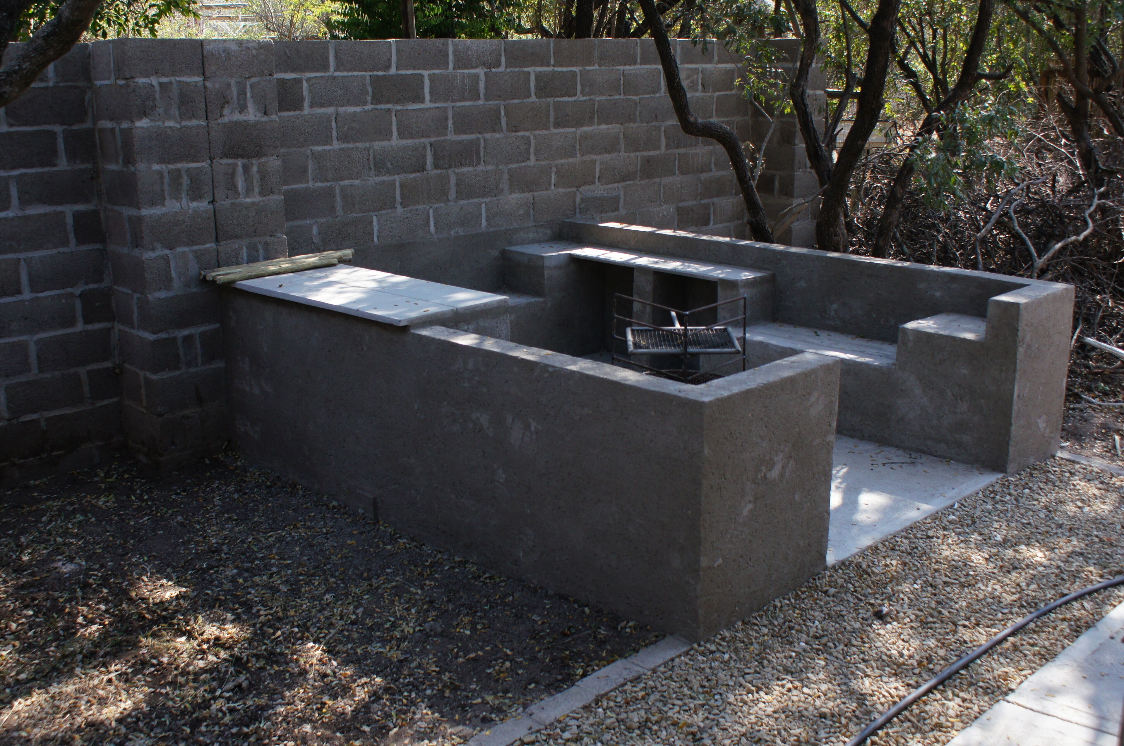 Ein Barbereich aus Beton mit einem Metallgrill, umgeben von einer Steinmauer und einem Kiesplatz.