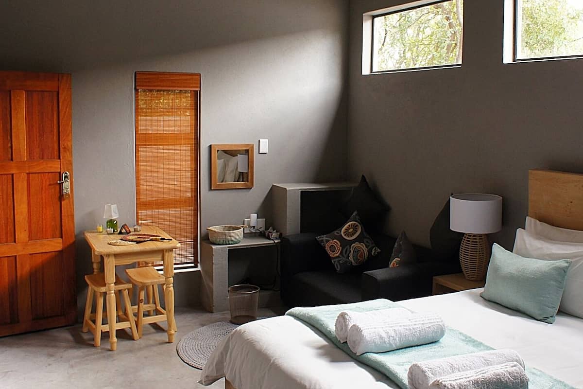 Ein Hotelzimmer mit Holztür, Schreibtisch mit Stuhl, Bett mit Kissen, Nachttisch mit Lampe und Fenster mit Blick auf Bäume.