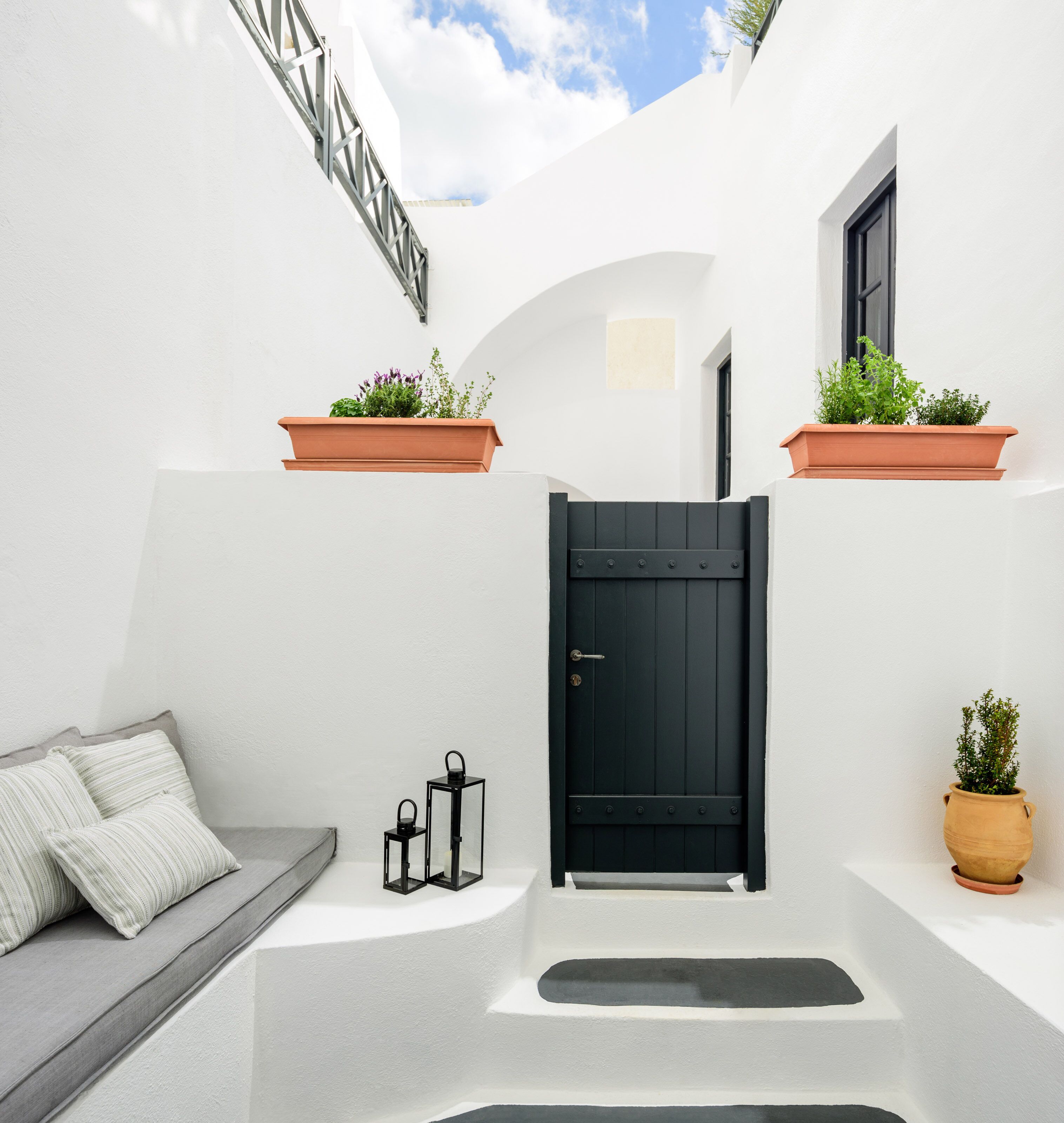 A white-walled interior with a black door, two potted plants, a bench with pillows, and two lanterns.