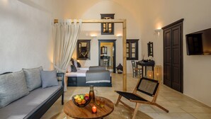 A cozy living room with a sofa, a coffee table with fruits and a candle, a wooden chair, and a bed in the background. - Firalia Suites (Santorini)