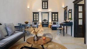 A spacious living room with a sofa, a coffee table, and a dining area with chairs and a table. - Firalia Suites (Santorini)