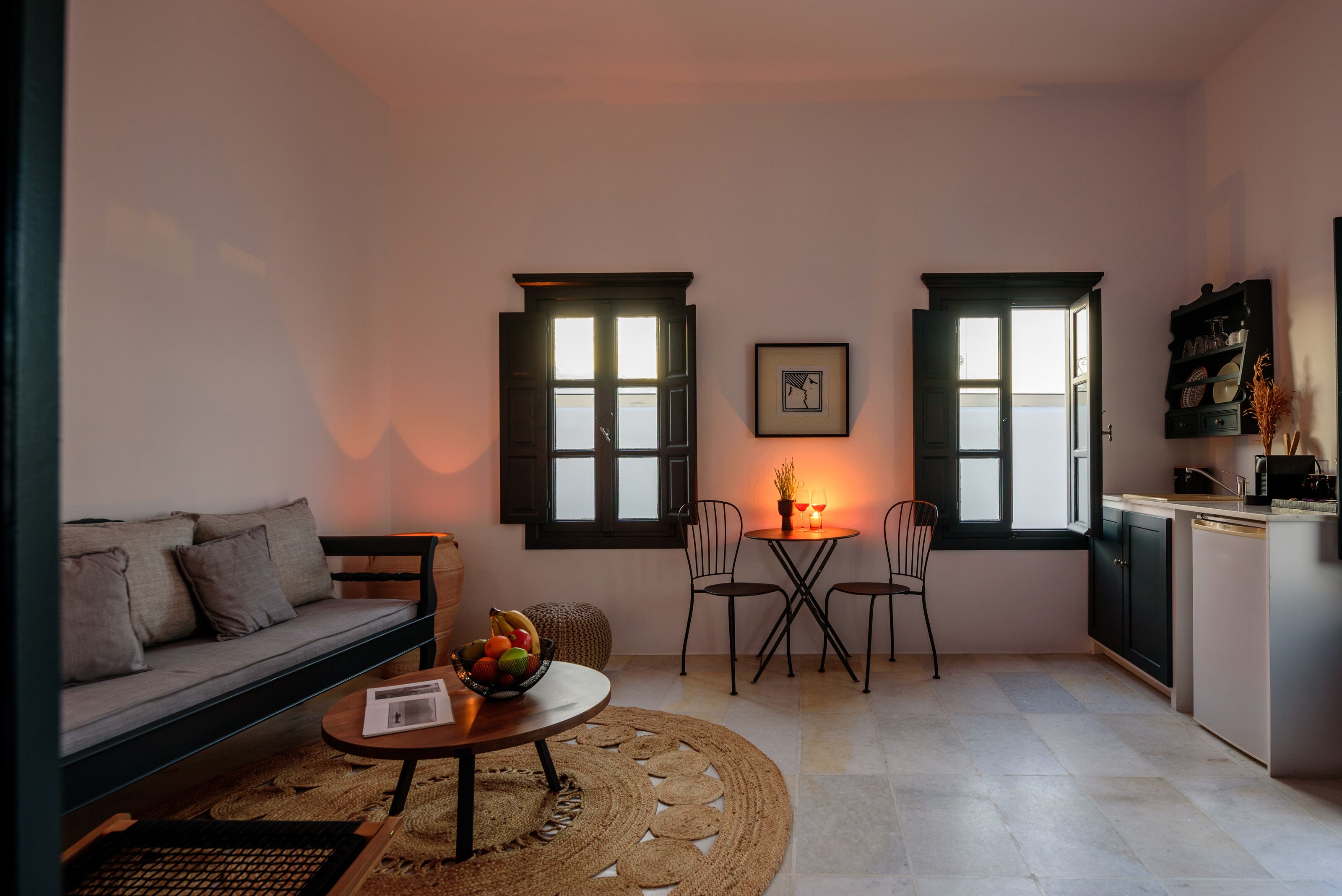 A compact living space with a sofa, a dining table with chairs, a small kitchen area, and two windows with shutters.