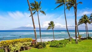 A tropical landscape with palm trees, a grassy area, and a view of the ocean. - Elegant Island-Inspired 2-Bedroom with Ocean Views (Lahaina)
