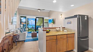A modern kitchen with wooden cabinets, a stainless steel refrigerator, and a view to the outside. - Elegant Island-Inspired 2-Bedroom with Ocean Views (Lahaina)