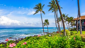 A beachfront property with palm trees, a view of the ocean, and a building with a balcony. - Elegant Island-Inspired 2-Bedroom with Ocean Views (Lahaina)
