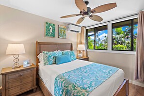 A bedroom with a bed, nightstands, a ceiling fan, a window with a view of greenery, and wall art. - Elegant Island-Inspired 2-Bedroom with Ocean Views (Lahaina)