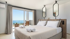 Ein Hotelzimmer mit einem großen Bett, einem Holzkopfende, zwei Spiegeln, Meerblick und einem Balkon mit Stuhl und Tisch.