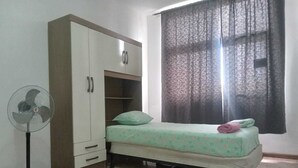 Un dormitori amb un llit, un armari, un ventilador de peu i una finestra amb cortines.