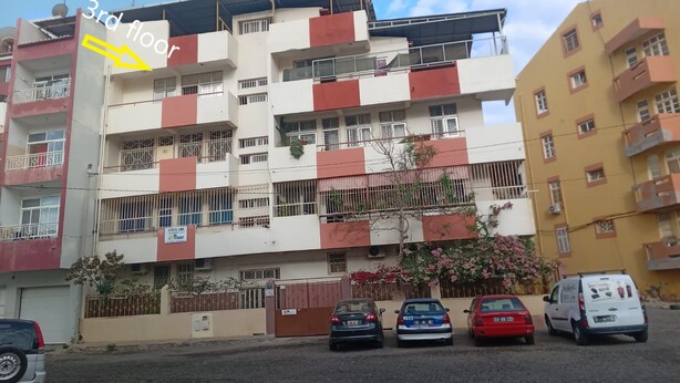 Un edifici residencial d'àmbit elevat amb balcons, una entrada tancada i cotxes aparcats.