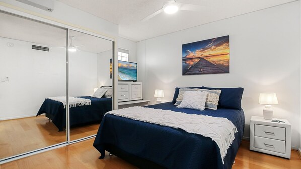 A bedroom with a large mirror, a bed with a blue bedspread, a nightstand with a lamp, and a painting of a sunset on the wall. - Grand Pacific, Unit 65, 100 Bulcock St, Caloundra (Caloundra)