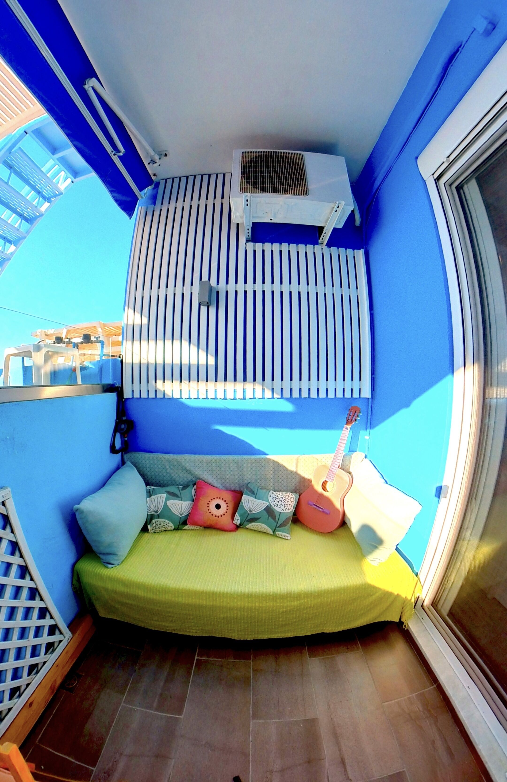 A small balcony with a green cushioned seating area, a white air conditioning unit, and a window with blinds.