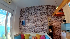 A bedroom with a curved bed, a stone wall, a small kitchen, and a balcony. - Beach Shack Studio s&l (Rhodes)