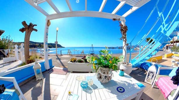 A covered outdoor seating area with a view of the sea, featuring a white pergola, blue and white furniture, and potted plants. - Beach Shack Studio s&l (Rhodes)