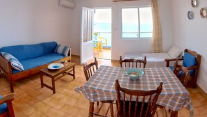 Ein geräumiges Wohnzimmer mit einem blauen Sofa, einem Holzespeisetisch mit kariertem Tischläufer und Meerblick durch große Fenster.