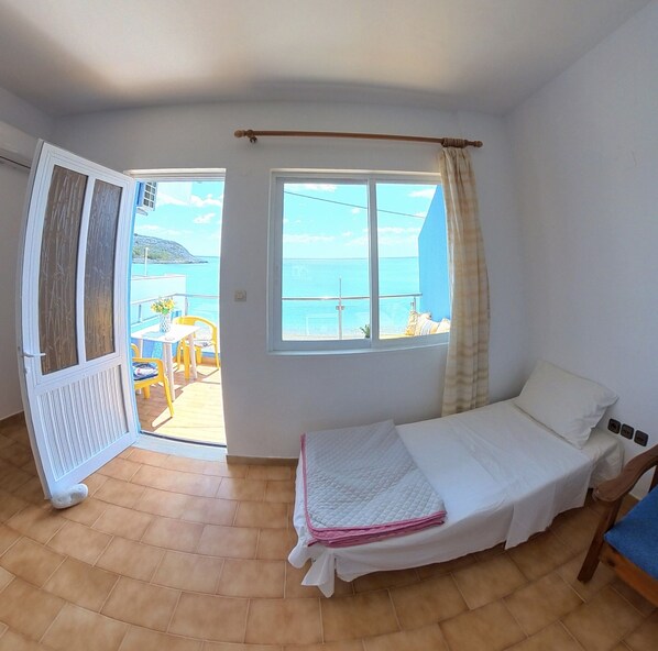 A bedroom with a bed, a window with a view of the sea, and a door leading to an outdoor area with a table and chairs.
