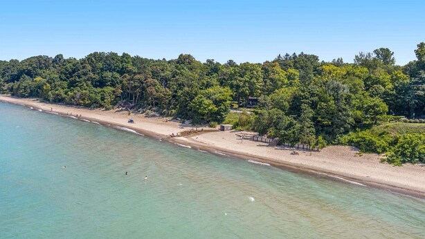 Eine Vogelperspektive zeigt einen Strand mit klarem Wasser, einem sandigen Ufer und dicht bewaldeten Hügeln im Hintergrund.