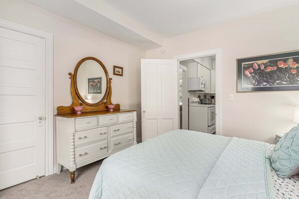 A bedroom with a bed, dresser, mirror, and a kitchen area visible through an open door.