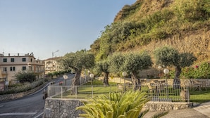 A hillside with a stone retaining wall, a paved road, and a fenced outdoor seating area with tables and chairs. - La Risacca Relais  (Belvedere Marittimo)