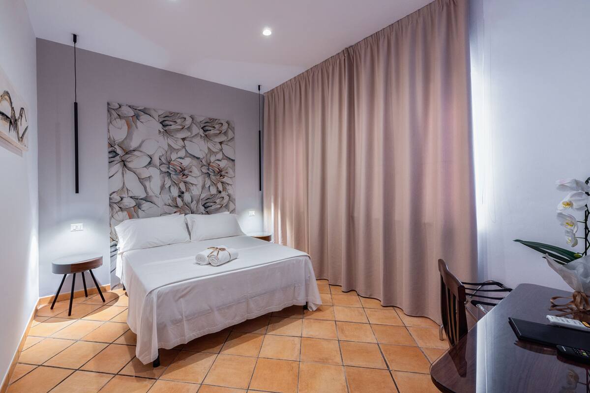 Un dormitorio con cama, mesita de noche, silla, escritorio y un ventanal con cortinas.