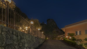 A nighttime view of a stone wall with a metal fence and street lamps, leading to a building with a sloped roof and a stone foundation. - La Risacca Relais  (Belvedere Marittimo)