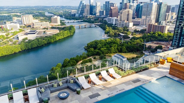Une piscine sur le toit, dotée de chaises longues et d’une balustrade en verre offrant une vue sur une rivière et le panorama de la ville.