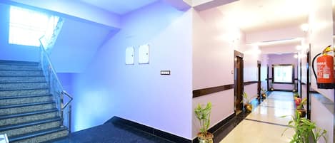 A hotel lobby with a staircase, potted plants, a fire extinguisher, and a reception area.