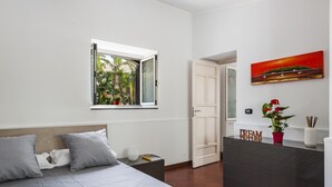 Quarto com cama, janela com vista para vegetação, pintura na parede e cômoda com uma planta.