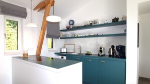 A modern kitchen with teal cabinetry, a central island, pendant lighting, and a window with a view of greenery. - Freistehender Bungalow in der Künstlerkolonie Ahrenshoop (Ostseebad Ahrenshoop)