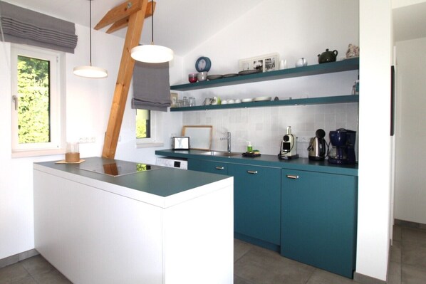 Une cuisine moderne avec des meubles de couleur turquoise, un îlot central, des luminaires suspendus et une fenêtre donnant sur un paysage verdoyant.