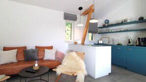 A modern living room with a sofa, a small round table, and a chair with a fur throw. A kitchen area with blue cabinets and a wooden hanging light fixture is visible. - Freistehender Bungalow in der Künstlerkolonie Ahrenshoop (Ostseebad Ahrenshoop)