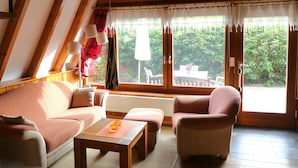 A cozy room with a sloped ceiling, a sofa, a chair, a table, and a view of greenery outside.