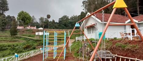 A playground with swings and climbing structures, surrounded by a white fence and houses in the background.