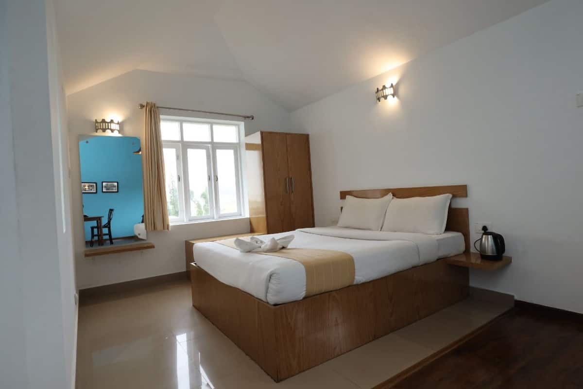 Een slaapkamer met een houten bed, een raam met gordijnen, een nachtkastje met waterkoker en een zithoek met een stoel en tafeltje.