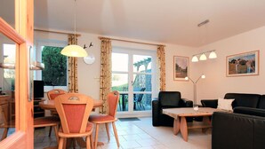 Sala de estar bem iluminada com área de jantar, sofá e mesa. Há cortinas nas janelas e um lustre no teto.