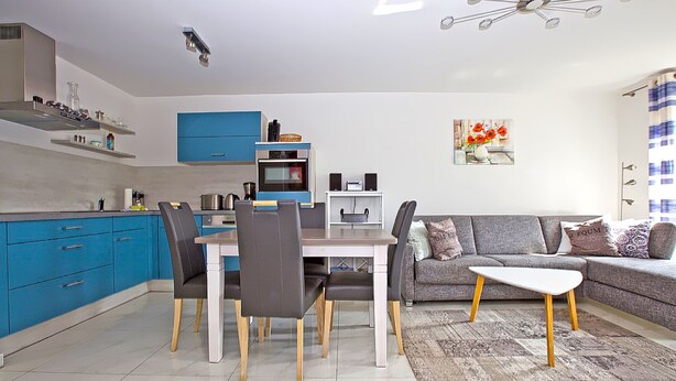 A modern kitchen with blue cabinets, a dining area with a white table and chairs, and a grey sofa with a patterned throw. - In Unmittelbarer Strandnähe ca. 150m (Ostseebad Dierhagen)