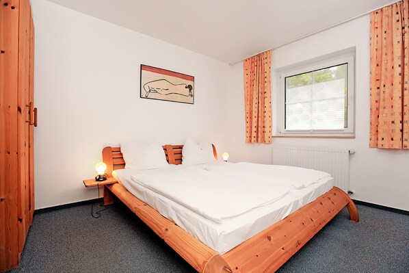 Ein Schlafzimmer mit einem hölzernen Bett, einem Nachttisch mit Lampe, einem Fenster mit Vorhängen und einem Wandbild.