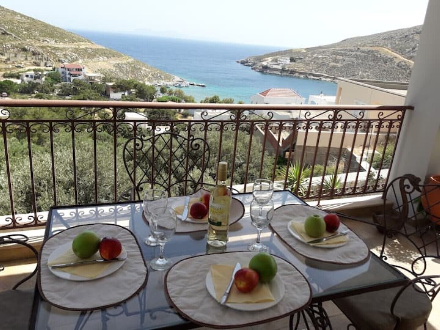 A table set for three with plates of fruit and glasses, overlooking a coastal landscape.