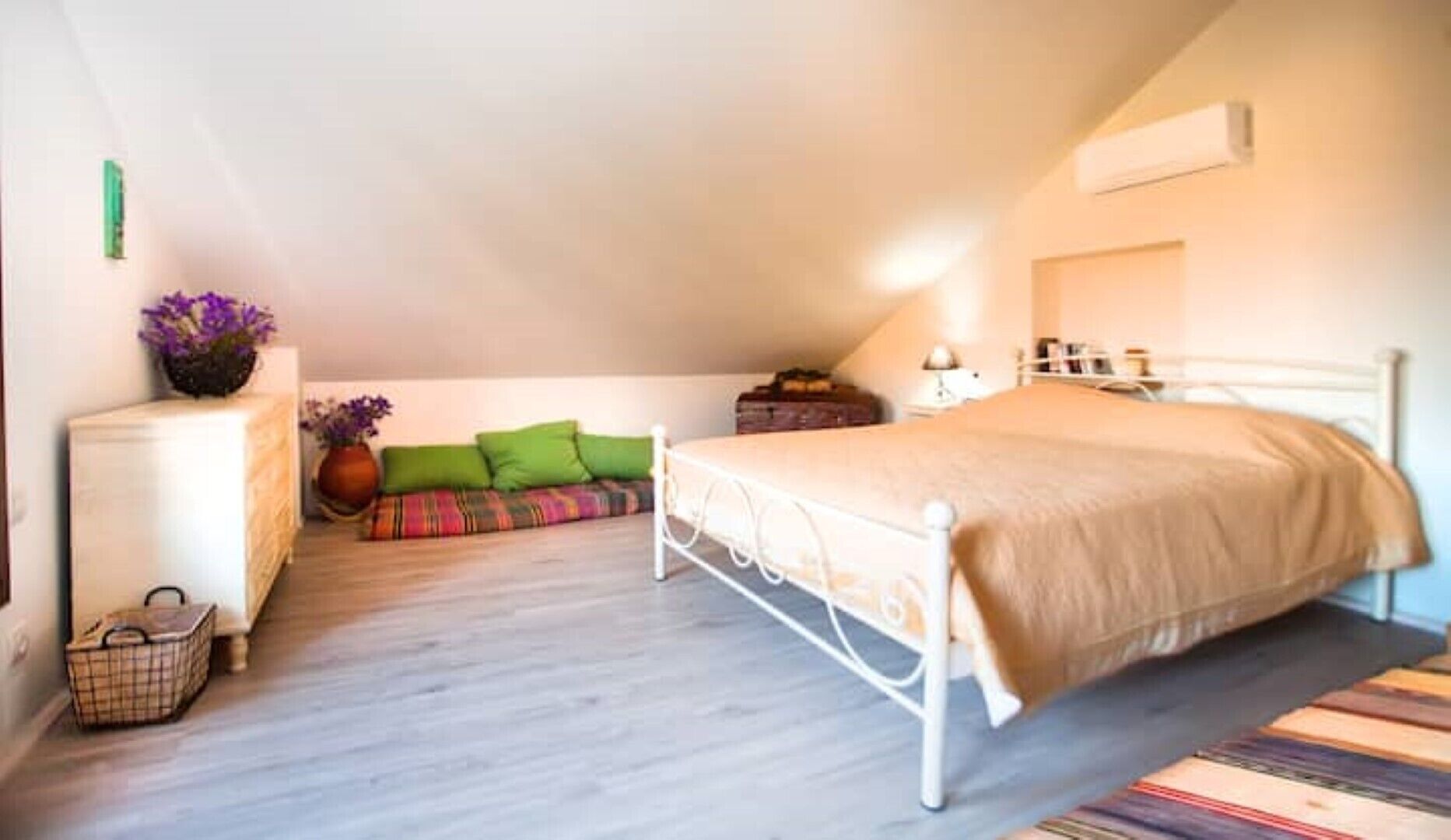 A bedroom with a white metal bed frame, a beige bedspread, a small bedside table with a lamp, and a colorful rug.