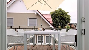 A balcony with a white table and chairs, a beige umbrella, and a view of a building with a red roof and a tree.