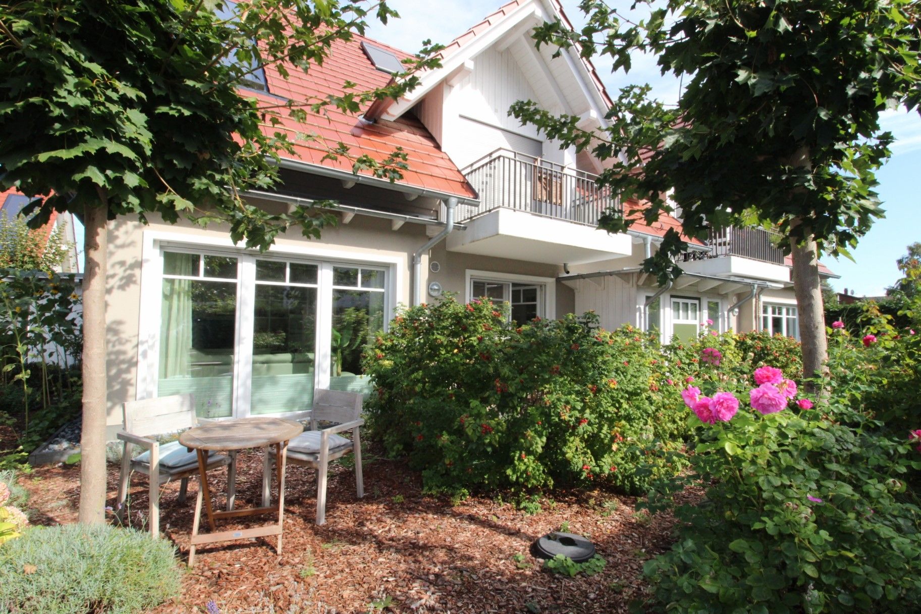 Ein zweigeschossiges Haus mit einem Balkon, einem Garten mit blühenden Pflanzen und einer Terrasse mit Tisch und Stühlen.