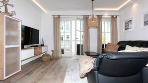 A modern living room with a black leather sofa, a flat-screen TV, wooden flooring, and a large window with curtains. - In Unmittelbarer Strandnähe ca. 100m (Ostseebad Wustrow)
