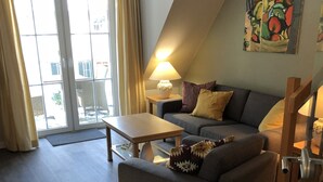 A modern living room with a sofa, a coffee table, and a lamp. There is a large window with curtains and a painting on the wall.