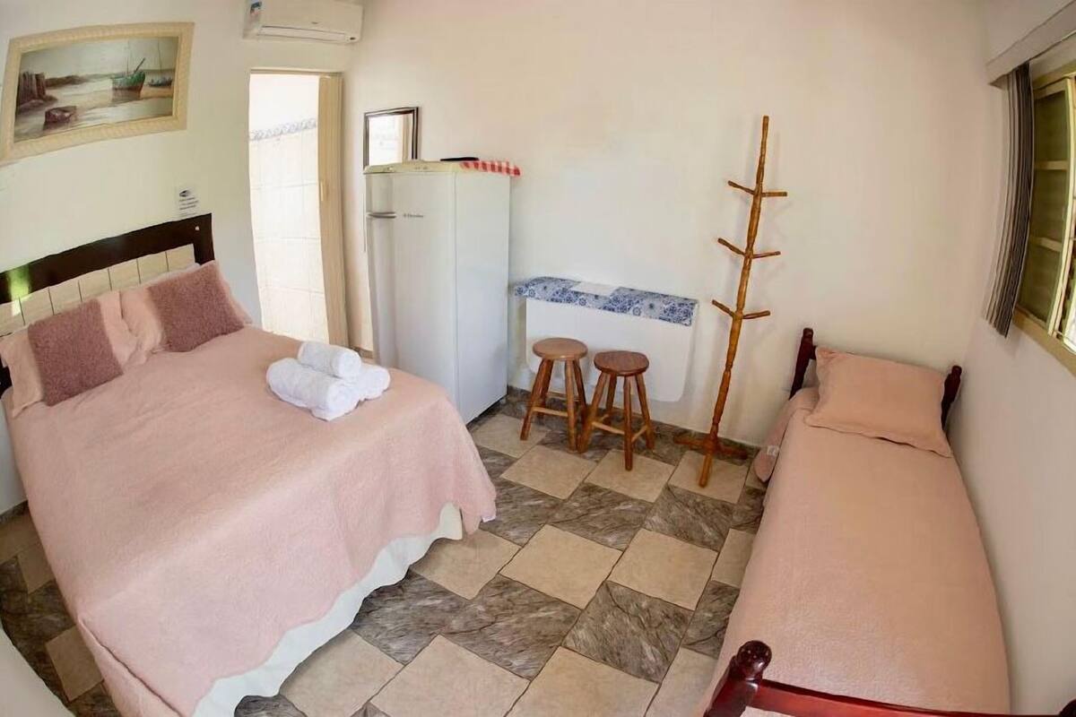 Un dormitorio con cama, una mesita, una silla, perchero y refrigerador.