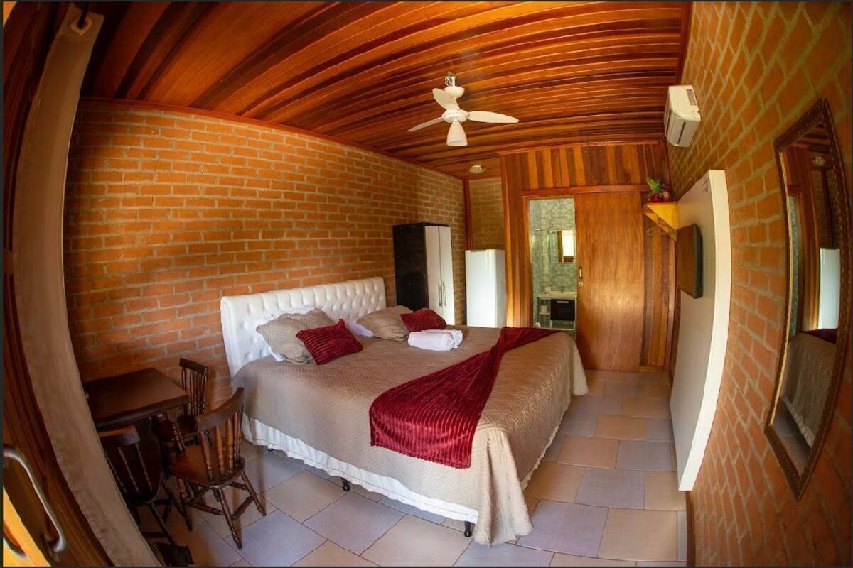 Un dormitorio con pared de ladrillo, techo de madera, cama con cabecera blanca, manta roja y ventilador de techo.