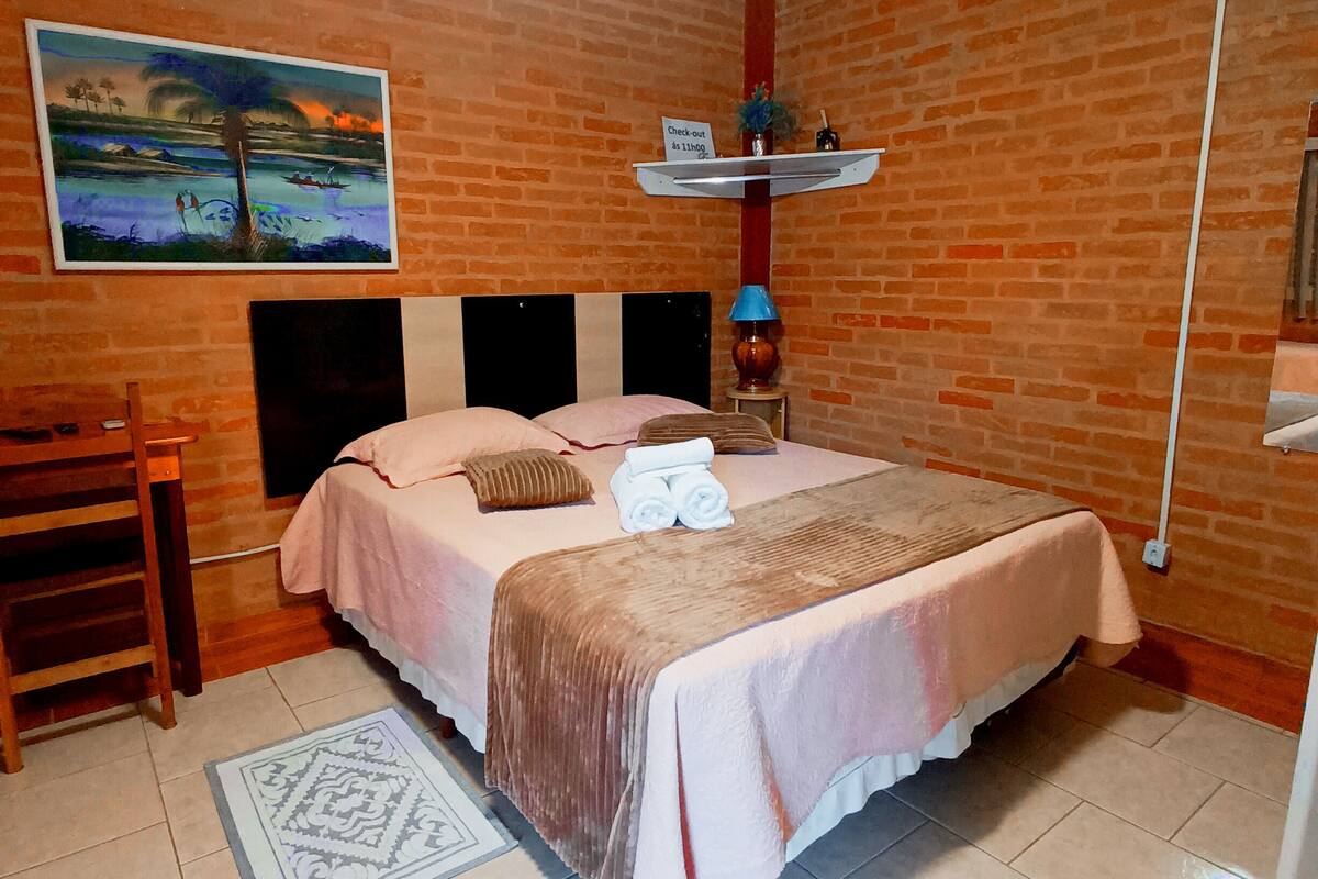 Habitación de hotel con pared de ladrillo, cama con dos almohadas, mesita de noche, un cuadro y un cartel de recepción.