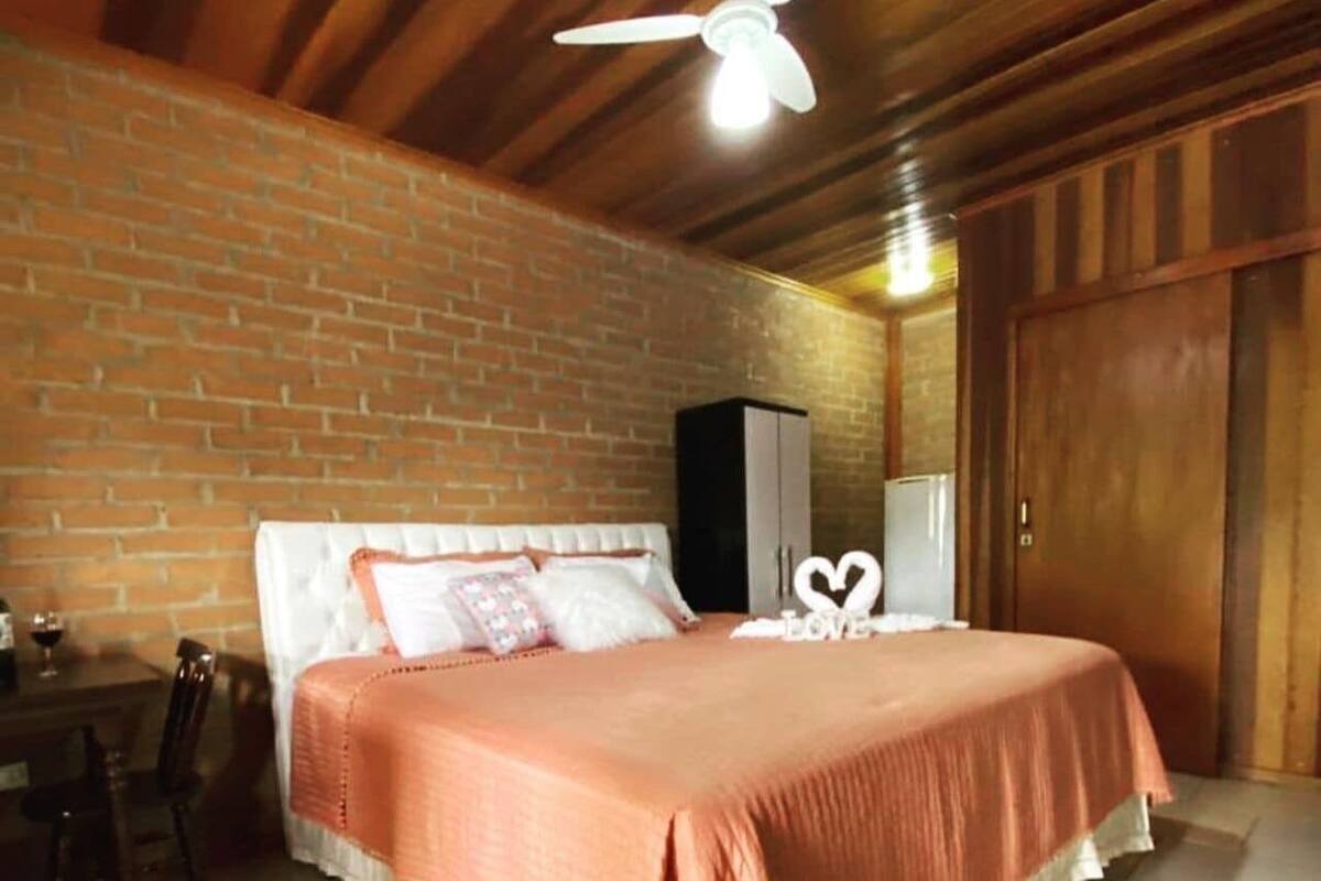 Un dormitorio con pared de ladrillo, cama con colcha rosa, ventilador de techo y armario.