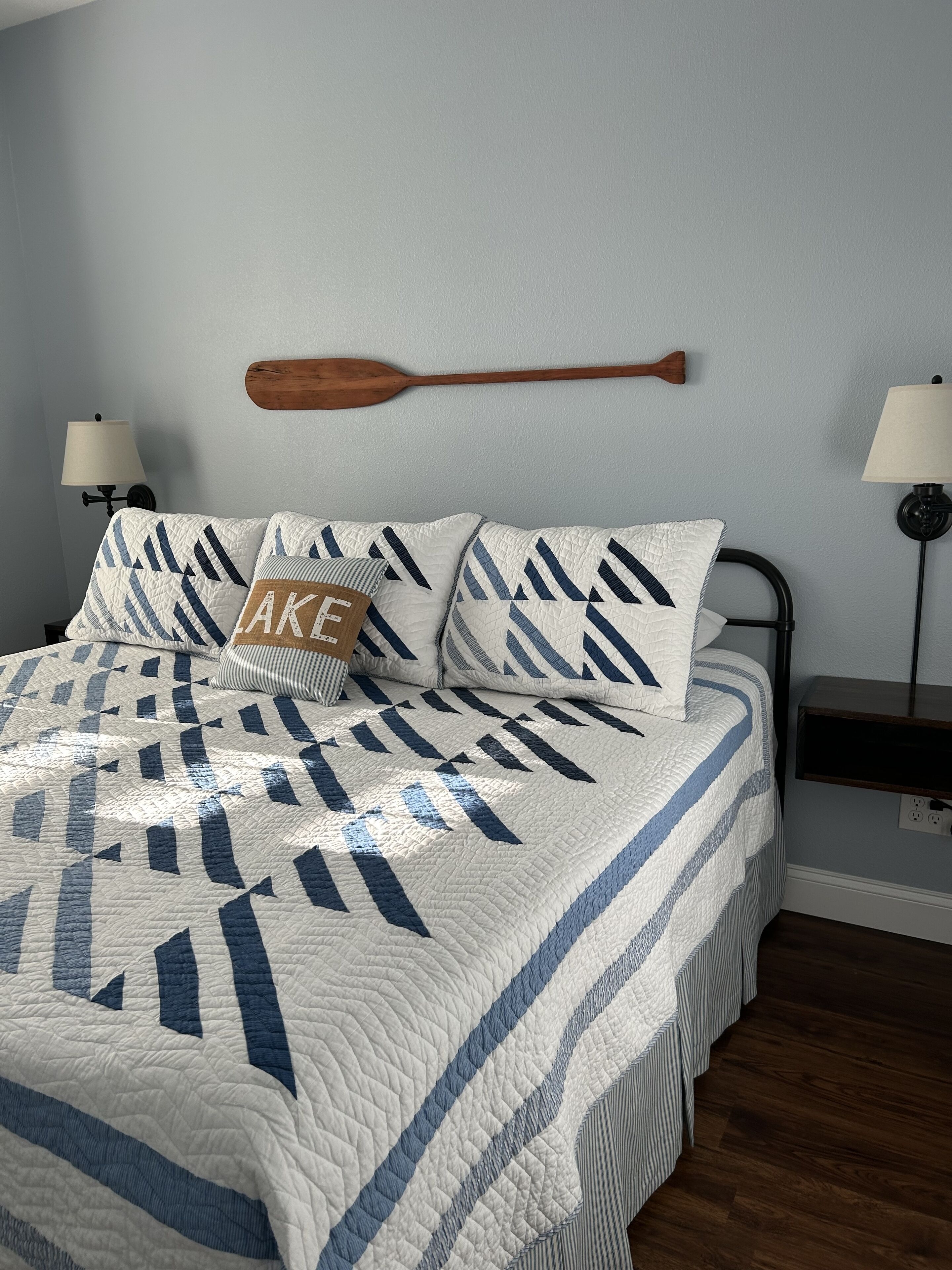 A neatly made bed with a quilt featuring a geometric pattern, a wooden paddle wall hanging, and two bedside lamps.