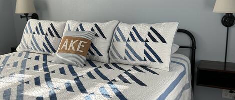 A neatly made bed with a quilt featuring a geometric pattern, a wooden paddle wall hanging, and two bedside lamps.