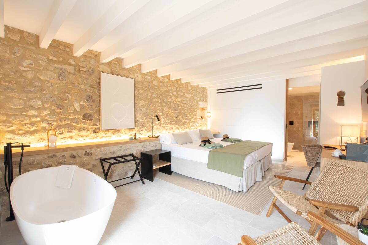 Un dormitorio moderno con pared de piedra, una bañera grande, una cama con manta verde y sillones de mimbre.